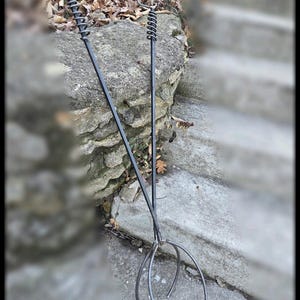 May include: Two black metal fire pit tools with spiral handles and curved tines. The tools are designed for handling logs and other items in a fire pit. The tools are handmade and have a rustic appearance.