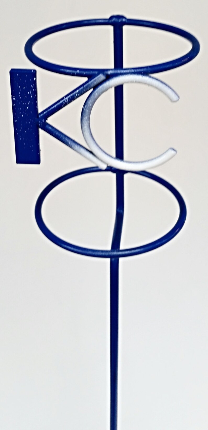 May include: A blue metal stand with two circular rings and a white and blue "KC" logo. The stand is designed to hold items such as glasses or bottles.