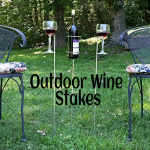 May include: Outdoor wine stakes with three metal poles, two wine glasses, and a bottle holder. The stakes are in a grassy area with two chairs in the background.