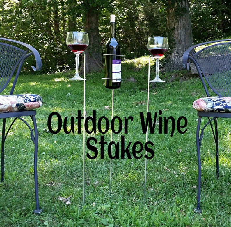 May include: Outdoor wine stakes with three metal poles, two wine glasses, and a bottle holder. The stakes are in a grassy area with two chairs in the background.