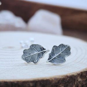 May include: A pair of silver-colored leaf-shaped stud earrings. The earrings are detailed with veining, and are displayed on a wooden surface. The earrings are a nature-inspired accessory.