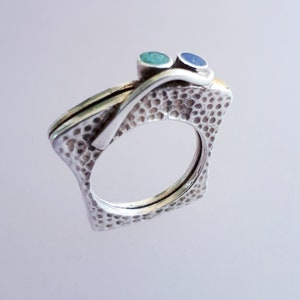Puede incluir: Un anillo de plata con un acabado texturizado y dos pequeñas piedras preciosas azules y verdes. El anillo tiene forma de cuadrado con esquinas redondeadas.