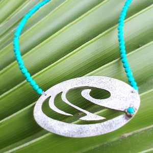 Puede incluir: Un collar de cuentas turquesa con un colgante de plata con un diseño de ola estilizado. El collar está sobre una hoja de palma verde.