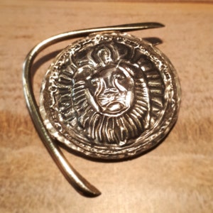 Könnte beinhalten: Eine goldfarbene Brosche aus Metall mit einem runden Design, das einen Löwenkopf in Relief zeigt. Die Brosche ist an einer gebogenen Metallstange befestigt.