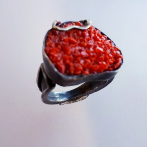 Puede incluir: Un anillo de plata con una gran piedra de coral rojo engastada en el centro. La piedra está rodeada por una banda de plata y tiene una superficie rugosa y texturizada.