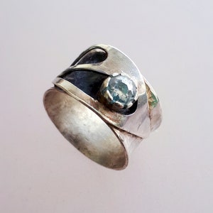 Può includere: Un anello d'argento con una pietra preziosa blu incastonata in una fascia testurizzata. L'anello è in metallo e ha un design unico.