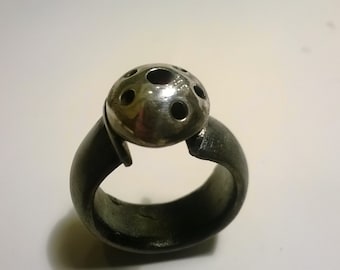 May include: A silver ring with a domed top featuring multiple circular cutouts. The ring band is made of a darker metal.