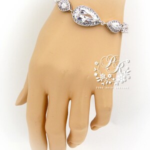 May include: A silver bracelet with a teardrop-shaped crystal and smaller crystals. The bracelet is on a mannequin's wrist.