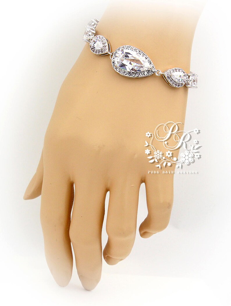 May include: A silver bracelet with a teardrop-shaped crystal and smaller crystals. The bracelet is on a mannequin's wrist.