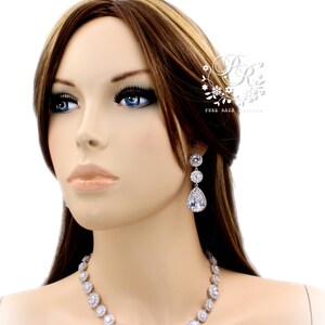 K&ouml;nnte beinhalten: Eine Frau tr&auml;gt eine silberne Halskette und Ohrringe mit tropfenf&ouml;rmigen Steinen. Die Ohrringe sind baumelnd und haben mehrere Steine. Die Halskette ist ein Choker-Stil mit runden Steinen.