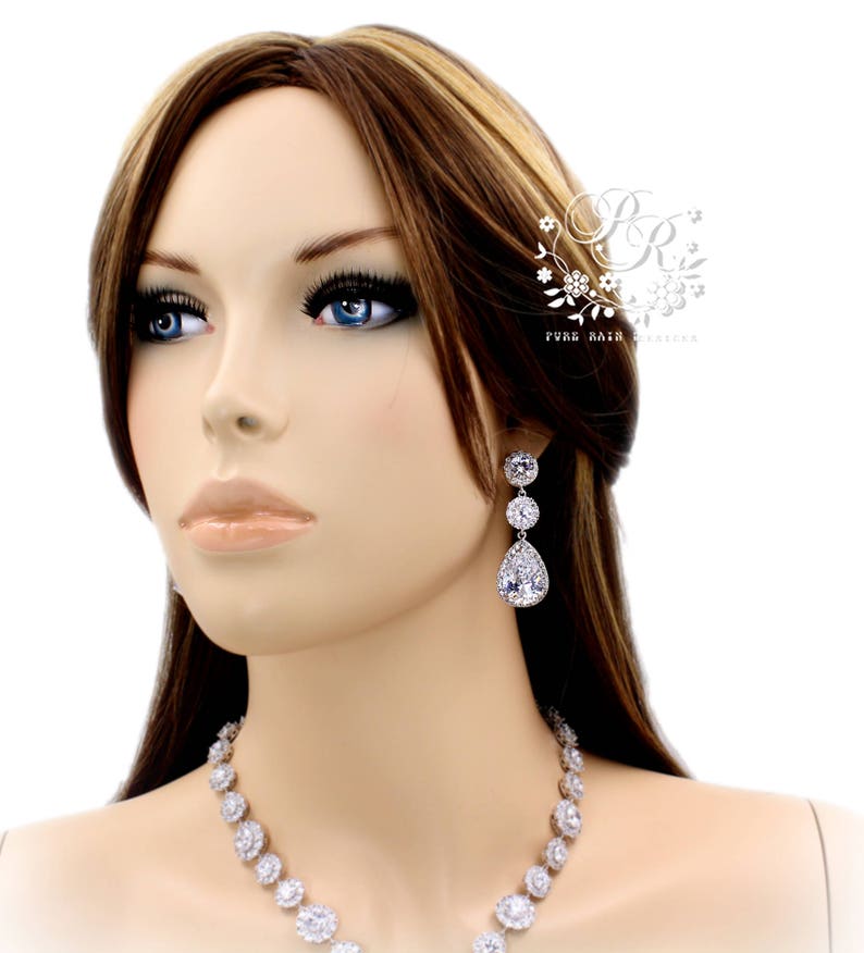 K&ouml;nnte beinhalten: Eine Frau tr&auml;gt eine silberne Halskette und Ohrringe mit tropfenf&ouml;rmigen Steinen. Die Ohrringe sind baumelnd und haben mehrere Steine. Die Halskette ist ein Choker-Stil mit runden Steinen.