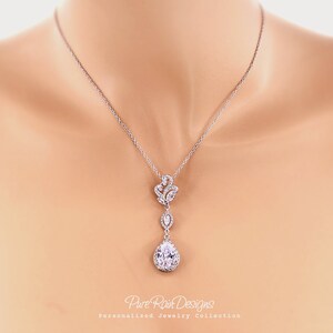 May include: A silver-toned pendant necklace featuring a teardrop-shaped, clear crystal pendant. The necklace has a delicate chain and a floral-inspired design above the teardrop. The text "Pure Rain Designs" and "Personalized Jewelry Collection" is visible.
