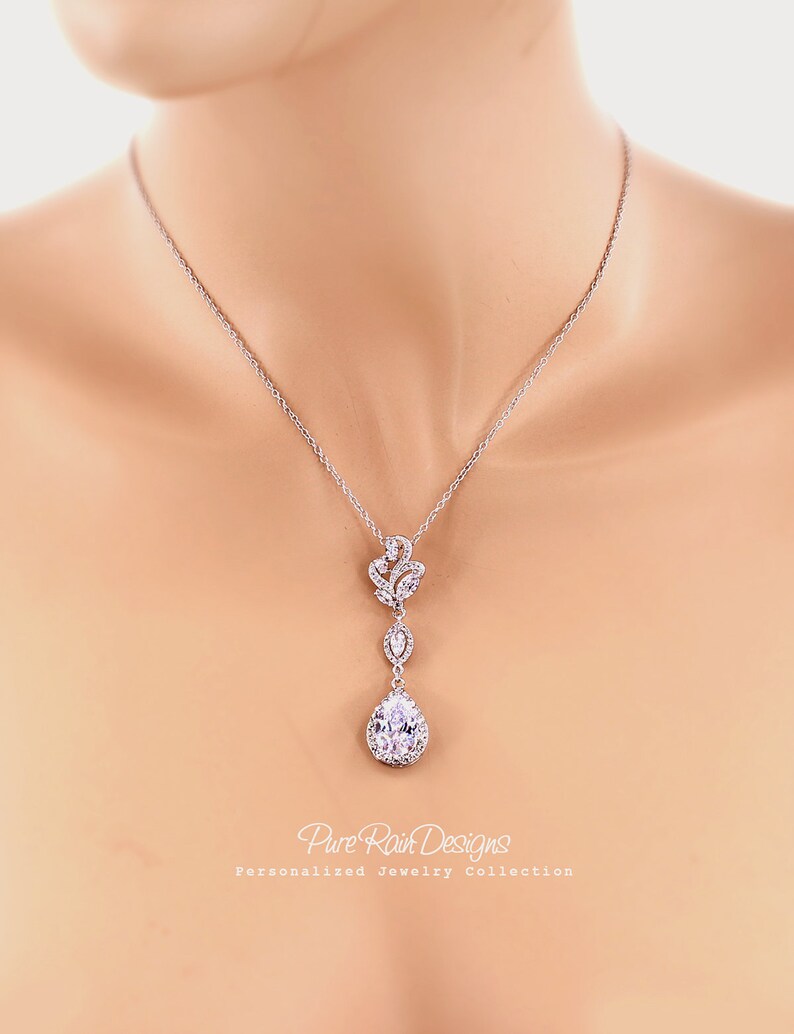May include: A silver-toned pendant necklace featuring a teardrop-shaped, clear crystal pendant. The necklace has a delicate chain and a floral-inspired design above the teardrop. The text "Pure Rain Designs" and "Personalized Jewelry Collection" is visible.