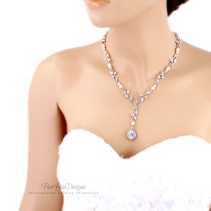 May include: A silver bridal necklace with a teardrop pendant featuring a large clear crystal surrounded by smaller crystals. The necklace is adorned with small pearl accents and delicate leaf designs.