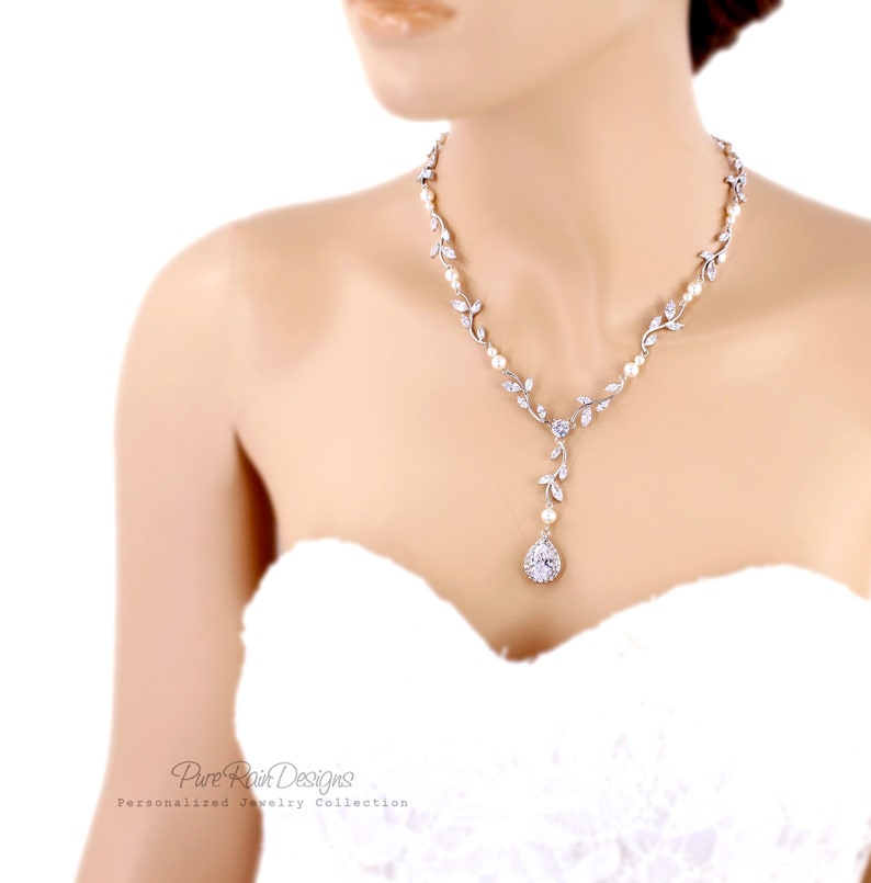 May include: A silver bridal necklace with a teardrop pendant featuring a large clear crystal surrounded by smaller crystals. The necklace is adorned with small pearl accents and delicate leaf designs.