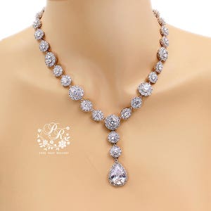K&ouml;nnte beinhalten: Eine silberne Halskette mit einem Tropfen-Anh&auml;nger, beide mit funkelnden, klaren Zirkonia-Steinen besetzt. Die Halskette hat eine zarte Kette und ein V-f&ouml;rmiges Design.