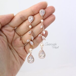 May include: Rose gold dangle earrings with pear-shaped crystals and a delicate leaf design. The earrings feature a pearl accent and are labeled "Swarovski Pearl".