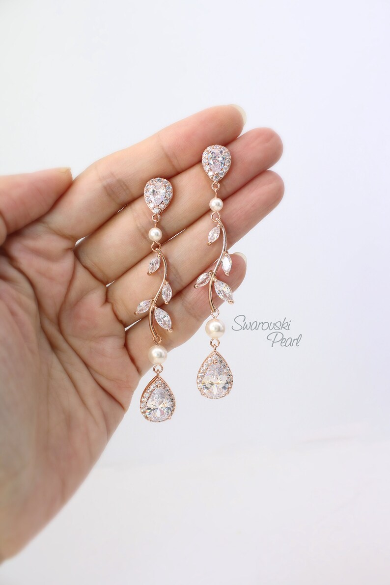 May include: Rose gold dangle earrings with pear-shaped crystals and a delicate leaf design. The earrings feature a pearl accent and are labeled "Swarovski Pearl".