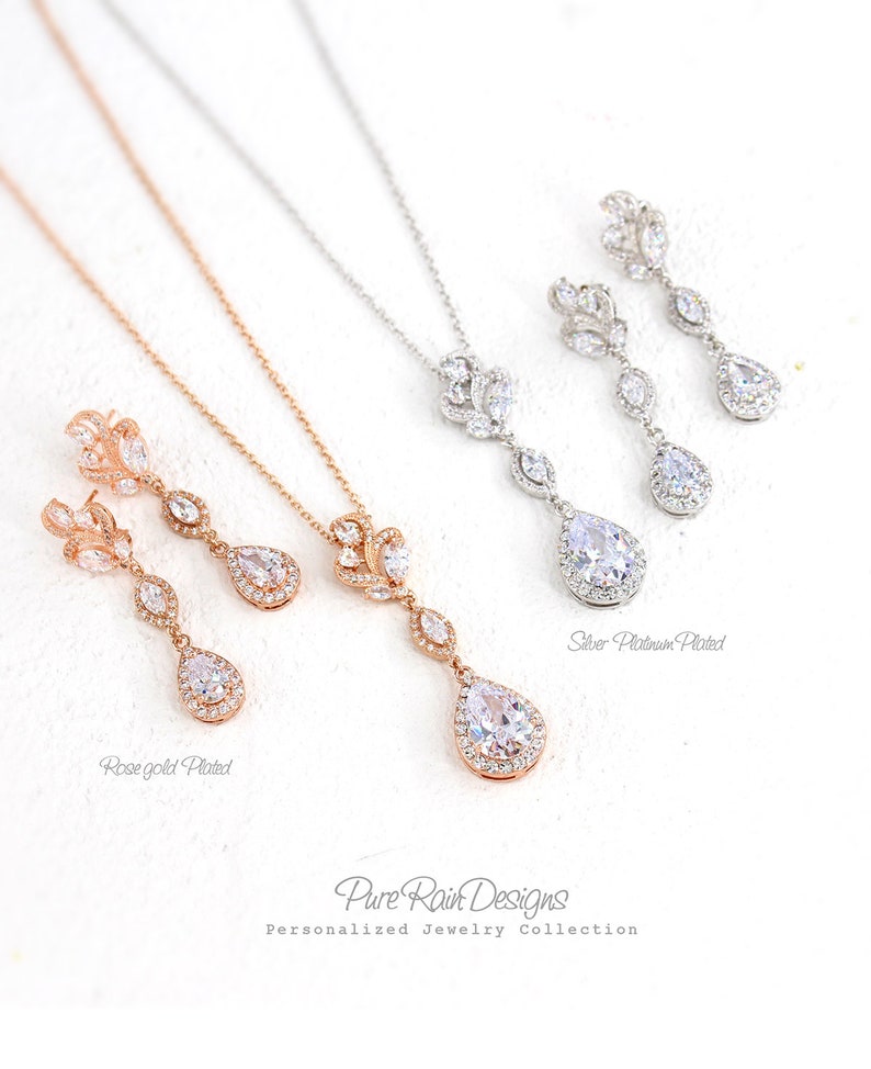 May include: Elegant jewelry sets featuring necklaces and matching earrings. The sets are available in rose gold and silver platinum plating. Each set includes a pendant necklace with a teardrop-shaped pendant and matching drop earrings. The jewelry is from Pure Rain Designs.