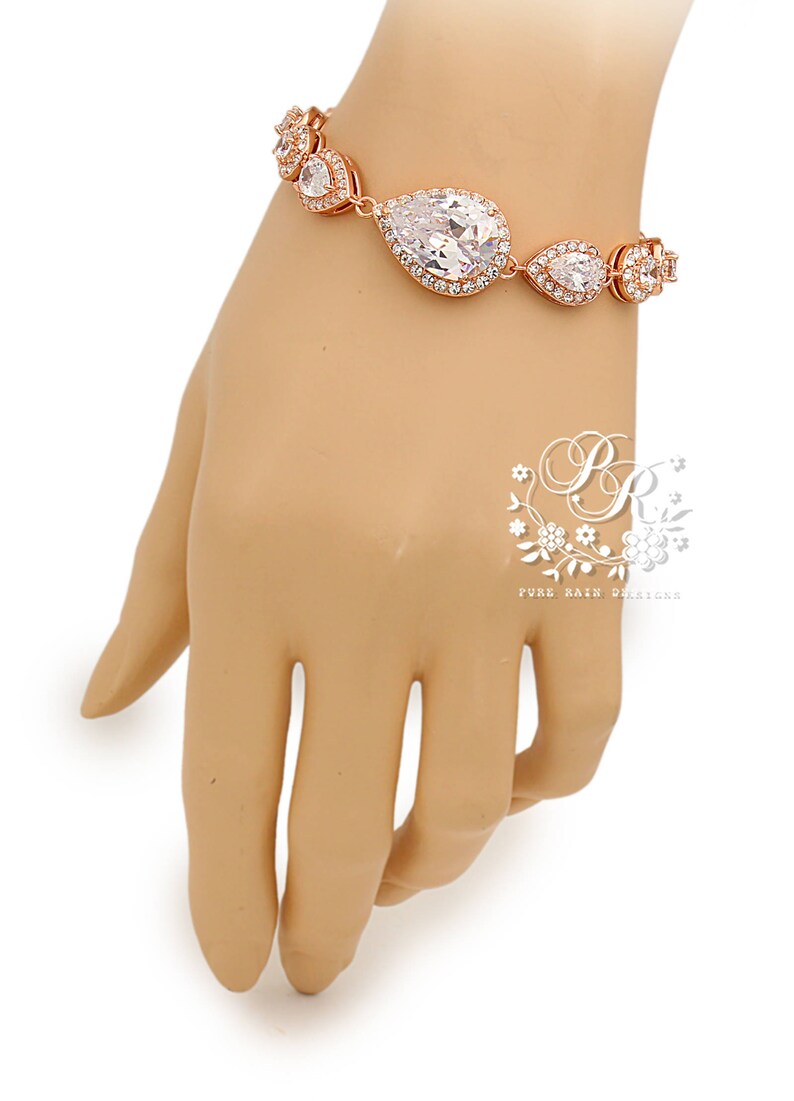 K&ouml;nnte beinhalten: Rosenvergoldetes Armband mit einem gro&szlig;en, klaren, birnenf&ouml;rmigen Kristall, der von kleineren Kristallen umgeben ist. Das Armband befindet sich an einer Mannequin-Hand.