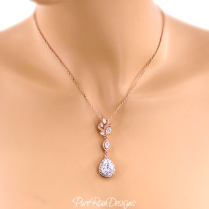 May include: Rose gold necklace with a pendant featuring a teardrop-shaped, clear crystal surrounded by smaller crystals. The necklace has a delicate chain and a floral-inspired design. The text "Pure Rain Designs" is visible.