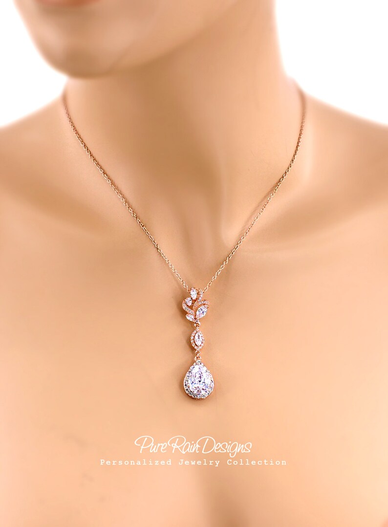 May include: Rose gold necklace with a pendant featuring a teardrop-shaped, clear crystal surrounded by smaller crystals. The necklace has a delicate chain and a floral-inspired design. The text "Pure Rain Designs" is visible.