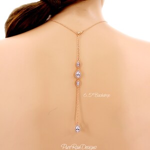 May include: A rose gold necklace with a 6.5-inch backdrop, featuring a series of clear, teardrop-shaped and oval-shaped crystals. The necklace is designed to be worn on the back, with a delicate chain and clasp.