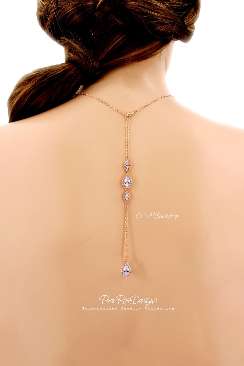May include: A rose gold necklace with a 6.5-inch backdrop, featuring a series of clear, teardrop-shaped and oval-shaped crystals. The necklace is designed to be worn on the back, with a delicate chain and clasp.