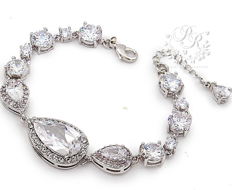 May include: A silver bracelet with a teardrop-shaped crystal pendant and smaller round crystals. The bracelet has a clasp closure.