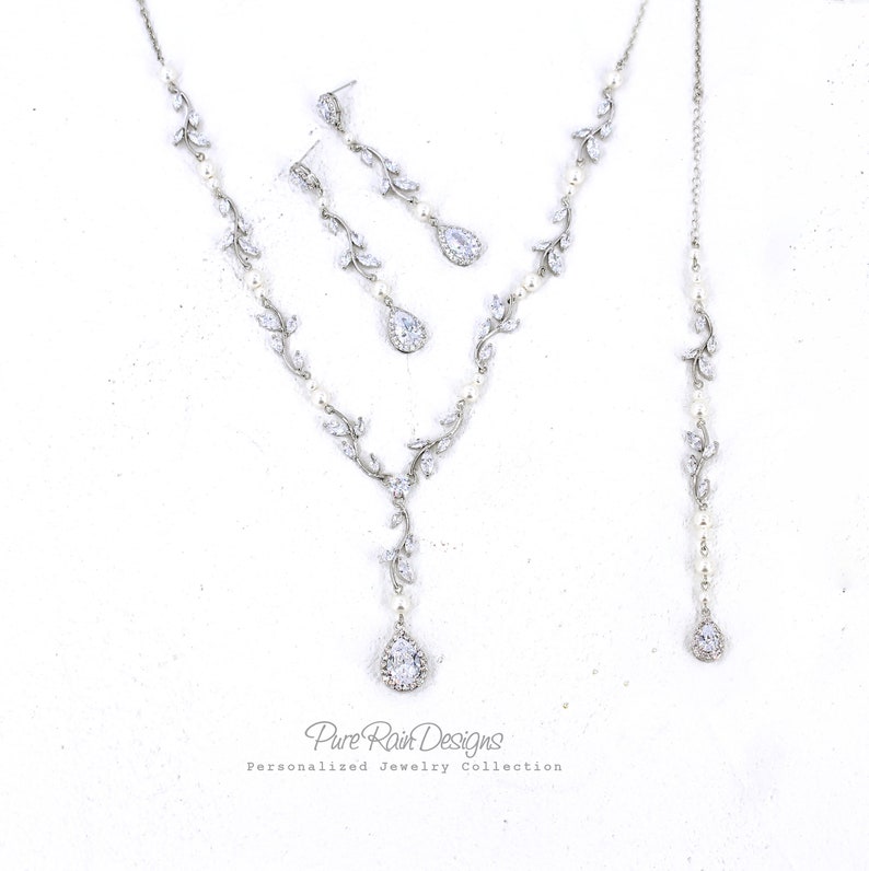 May include: A silver bridal jewelry set with a teardrop pendant necklace, matching earrings, and a pearl and crystal bracelet. The necklace features a delicate vine design with clear crystals and pearls.