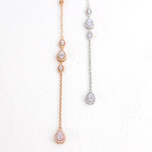 May include: Two delicate necklaces with a drop pendant design. One is rose gold, the other silver. Each features a chain with three graduated, sparkling teardrop-shaped stones and a larger teardrop pendant. The necklaces are displayed against a white background.
