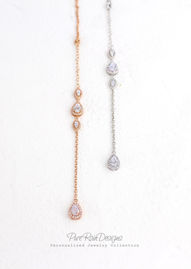 May include: Two delicate necklaces with a drop pendant design. One is rose gold, the other silver. Each features a chain with three graduated, sparkling teardrop-shaped stones and a larger teardrop pendant. The necklaces are displayed against a white background.