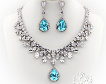 May include: A jewelry set consisting of a necklace and matching drop earrings. The necklace features a central light blue pear-shaped stone surrounded by clear crystals, with additional clear crystal accents along the silver-toned chain.