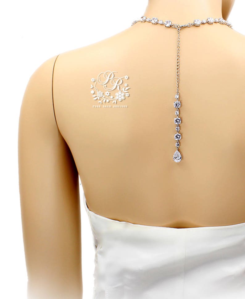 May include: A white bridal back necklace with a teardrop pendant and a small floral design with the initials "BR" on the back of a mannequin.
