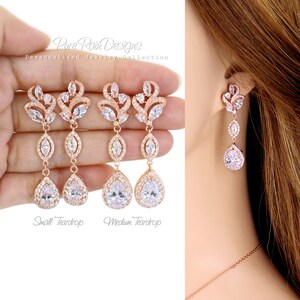 May include: Rose gold-toned drop earrings with clear, sparkling crystals. The earrings feature a floral design at the top, a marquise-shaped crystal in the middle, and a large teardrop-shaped crystal at the bottom. The text "Pure Rain Designs" is visible.