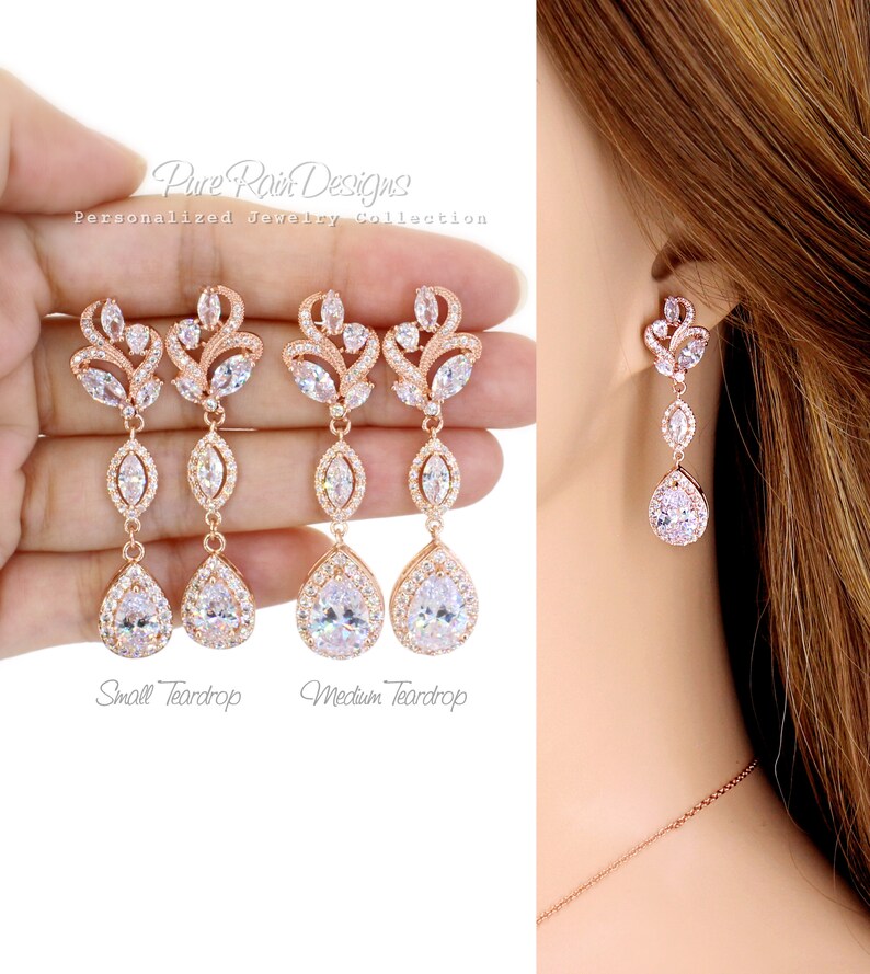 May include: Rose gold-toned drop earrings with clear, sparkling crystals. The earrings feature a floral design at the top, a marquise-shaped crystal in the middle, and a large teardrop-shaped crystal at the bottom. The text "Pure Rain Designs" is visible.