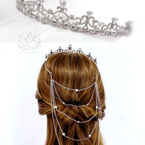 May include: Silver tiara with a delicate design and a chain of pearls hanging down the back. The tiara is worn on a model with long brown hair.