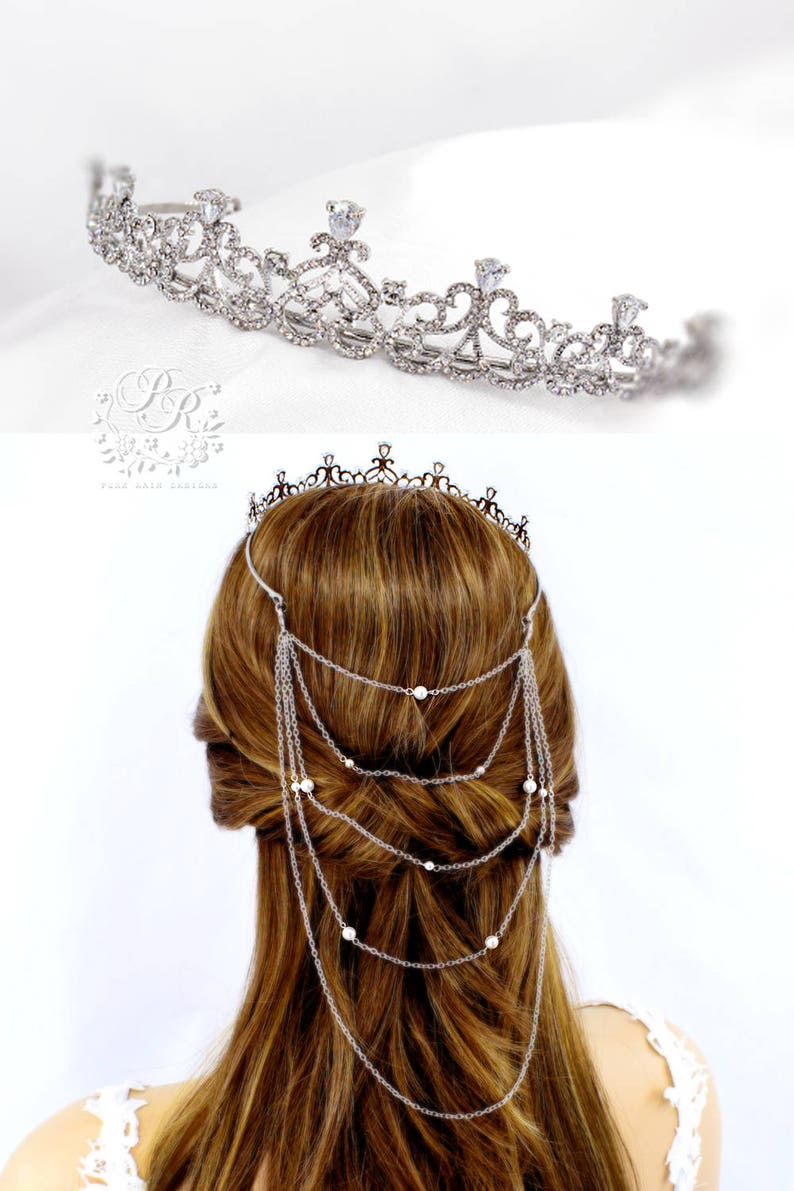 May include: Silver tiara with a delicate design and a chain of pearls hanging down the back. The tiara is worn on a model with long brown hair.