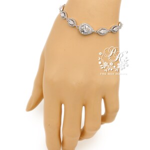 May include: A silver bracelet with clear, sparkling crystals. The bracelet features a central pear-shaped crystal surrounded by smaller, marquise-cut crystals. The bracelet is displayed on a mannequin hand against a white background.