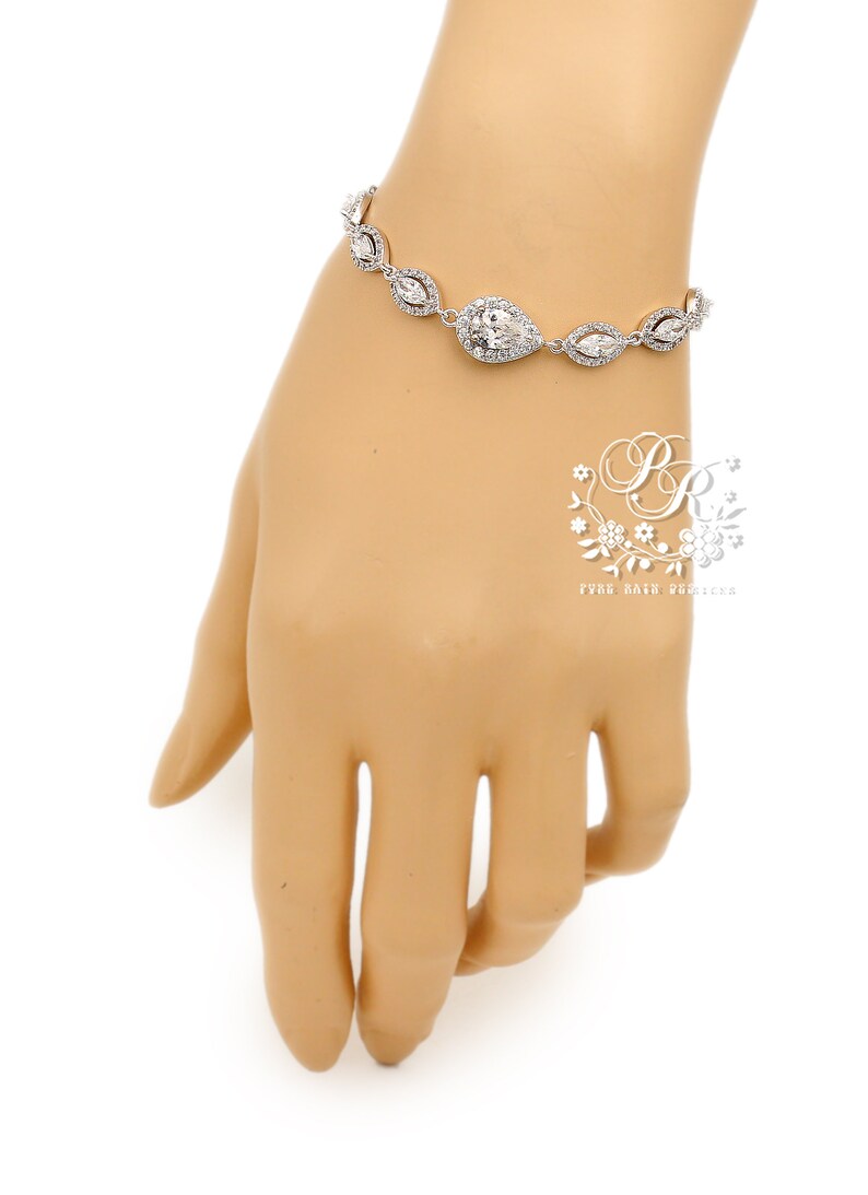 May include: A silver bracelet with clear, sparkling crystals. The bracelet features a central pear-shaped crystal surrounded by smaller, marquise-cut crystals. The bracelet is displayed on a mannequin hand against a white background.