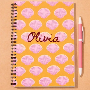Puede incluir: Cuaderno de espiral con un patrón repetido de conchas rosas sobre un fondo amarillo. El nombre "Olivia" está escrito en cursiva. Un bolígrafo rosa está colocado junto al cuaderno.