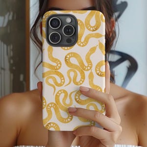 Peut inclure: Une coque de téléphone blanche avec un motif de serpent doré. Les serpents sont stylisés et ont un motif de croissant de lune sur leur corps.