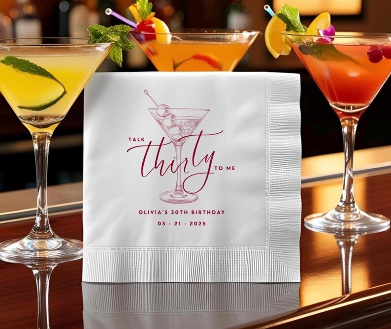 May include: A white square paper napkin featuring a maroon line drawing of a martini glass with olives. The text reads "TALK thirty TO ME" in a script font, followed by "OLIVIA'S 30TH BIRTHDAY" and the date "03 21 2025" in a clean sans-serif font.