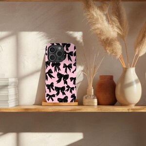 May include: A pink phone case with a black bow pattern. The case is designed for a smartphone with a camera bump.