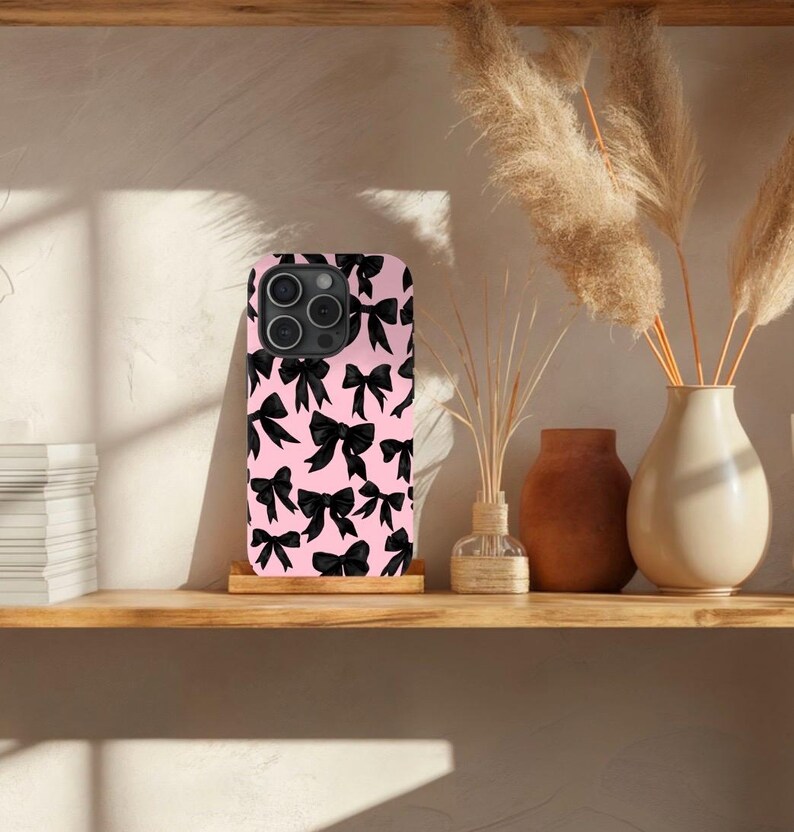 May include: A pink phone case with a black bow pattern. The case is designed for a smartphone with a camera bump.