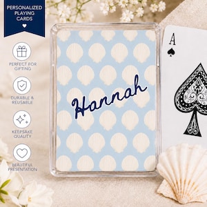 May include: A deck of personalized playing cards with a light blue background and a pattern of white seashells. The name "Hannah" is written in navy blue script across the center. An Ace of Spades card is visible.
