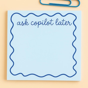 May include: A light blue notepad with a wavy dark blue border and the text "ask copilot later". A blue paperclip rests above the notepad on a light orange background. Stationery.