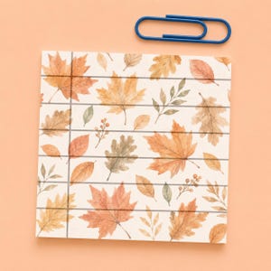 May include: A square notepad with a watercolor pattern of orange, yellow, and brown autumn leaves and small branches, featuring thin horizontal and vertical dark gray lines, accompanied by a single blue metal paperclip.