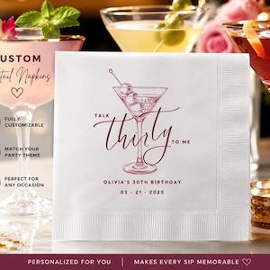May include: A white square cocktail napkin featuring a maroon graphic of a martini glass with olives and the text Talk thirty to me, Olivia's 30th Birthday, and 03 21 2025. The napkin has a textured border and is displayed in a party setting.