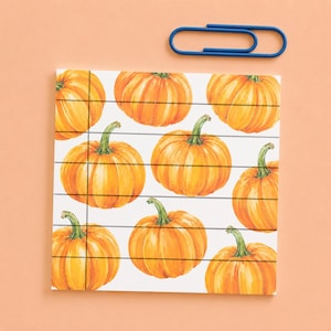 May include: A square notepad featuring a repeating watercolour pattern of orange pumpkins with green stems on a white background, overlaid with thin horizontal and vertical black lines, accompanied by a single blue metal paperclip.
